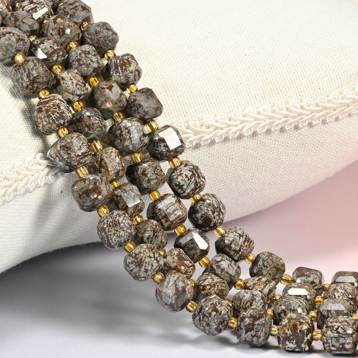 Brown snowflake obsidian faceted cube beads 8mm with small gold spacer beads, several strands draped over a cream cushion with braided trim