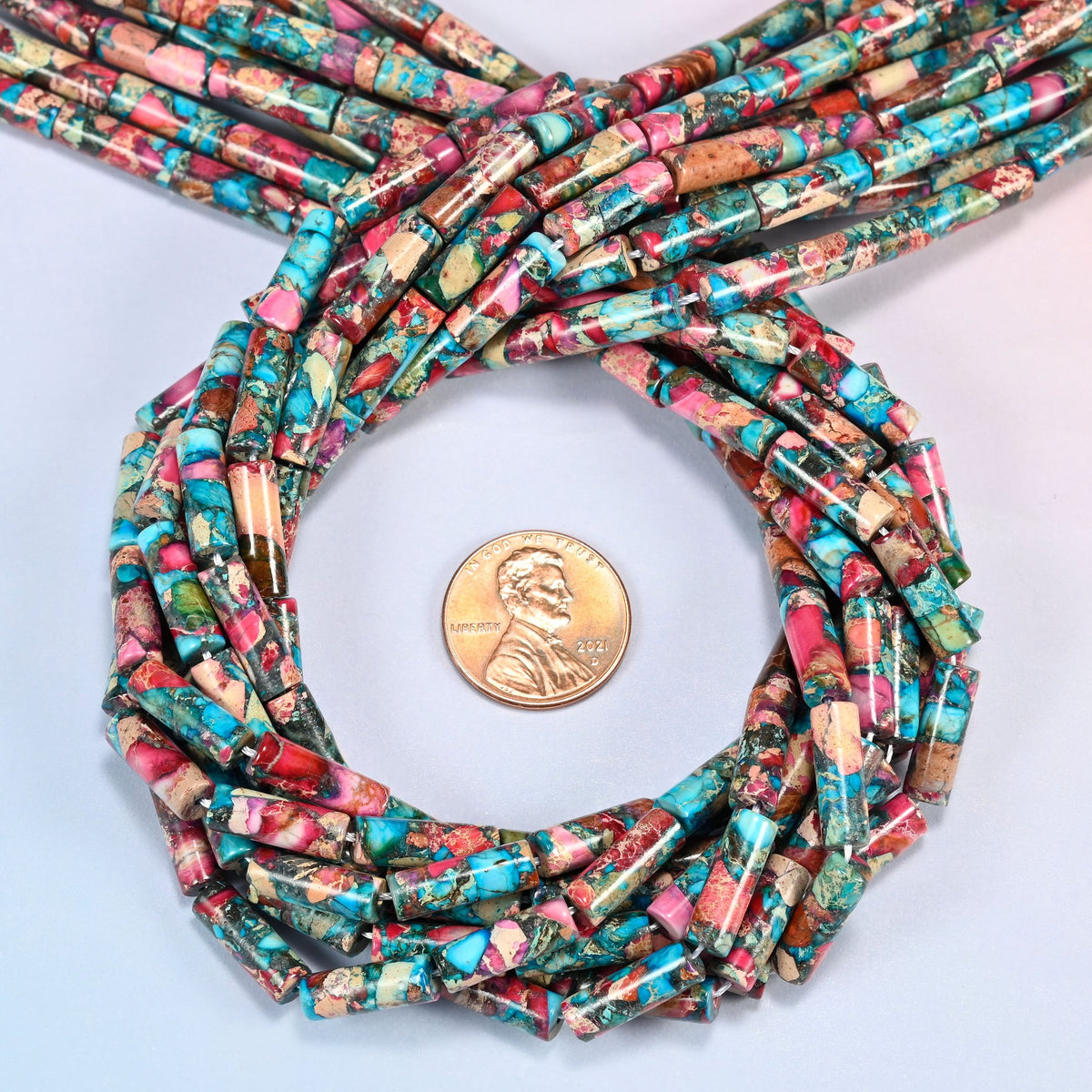 Dyed sea sediment jasper tube beads 13x4mm coiled in a loop around a US penny for scale, close-up of the slim tube shape