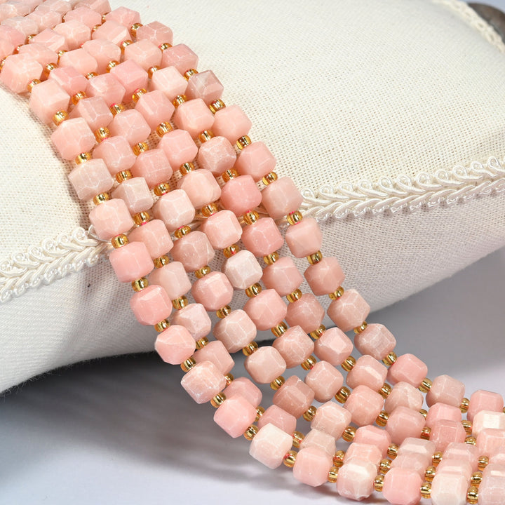 Natural pink opal faceted cube beads draped over a white linen cushion, salmon pink stone with gold glass seed bead spacers.