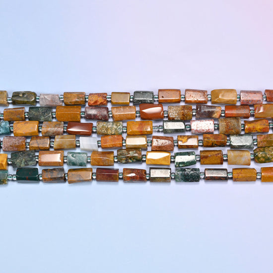 Overhead view of several rows of faceted Ocean Jasper tube beads in mustard, red, and green tones on a light lavender surface.