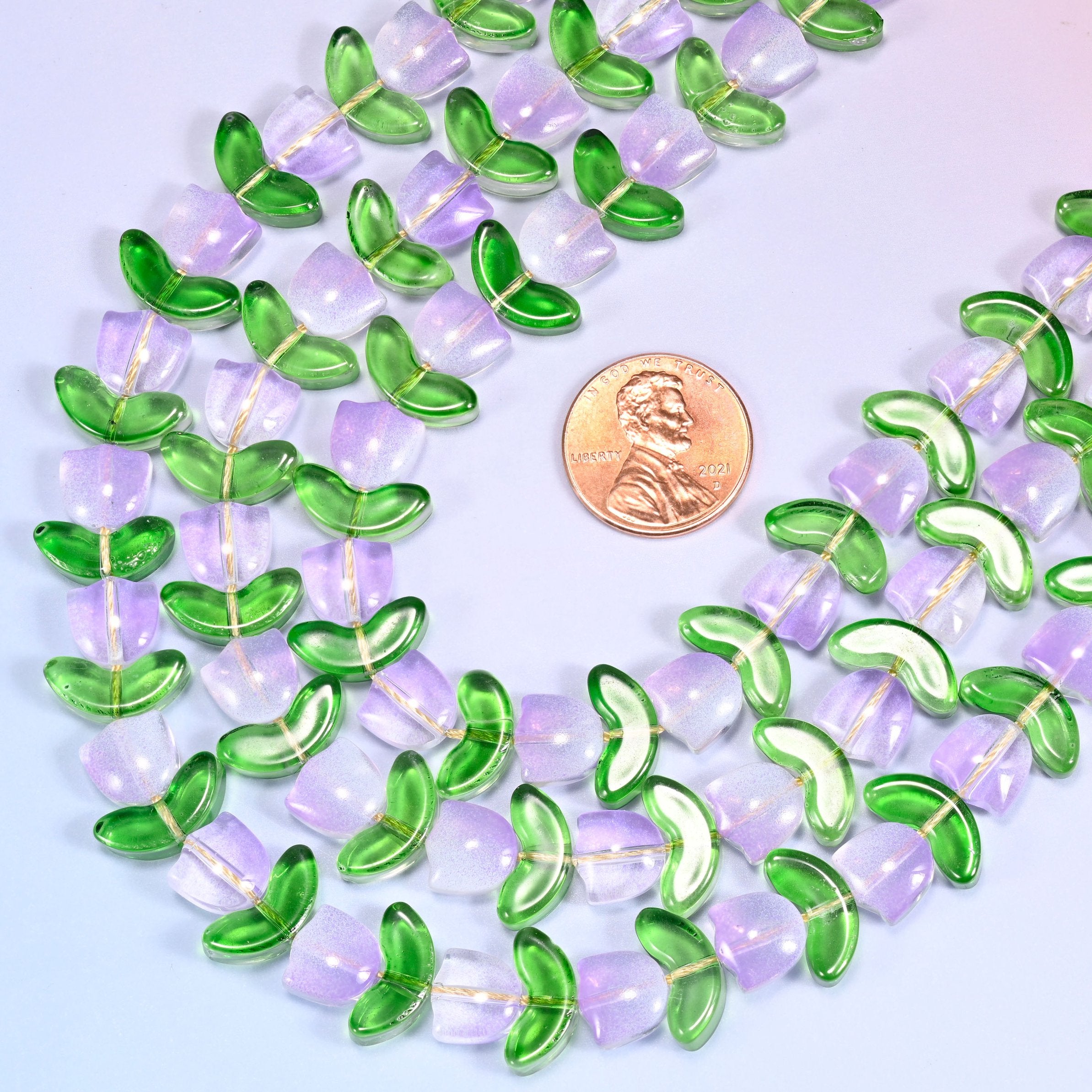Circular coil of lavender tulip and green leaf glass bead strands forming a wreath shape on a lavender background.