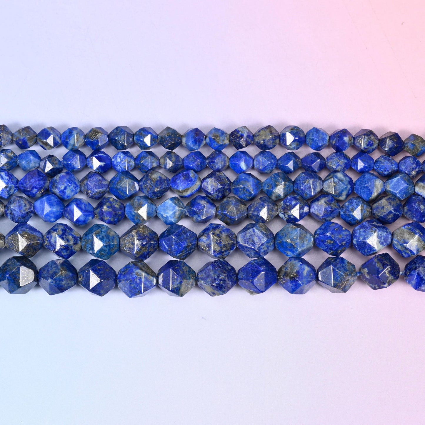 Blue Lapis Lazuli faceted beads in flat horizontal rows on a soft pink and lavender background, sold per 15.5 inch strand