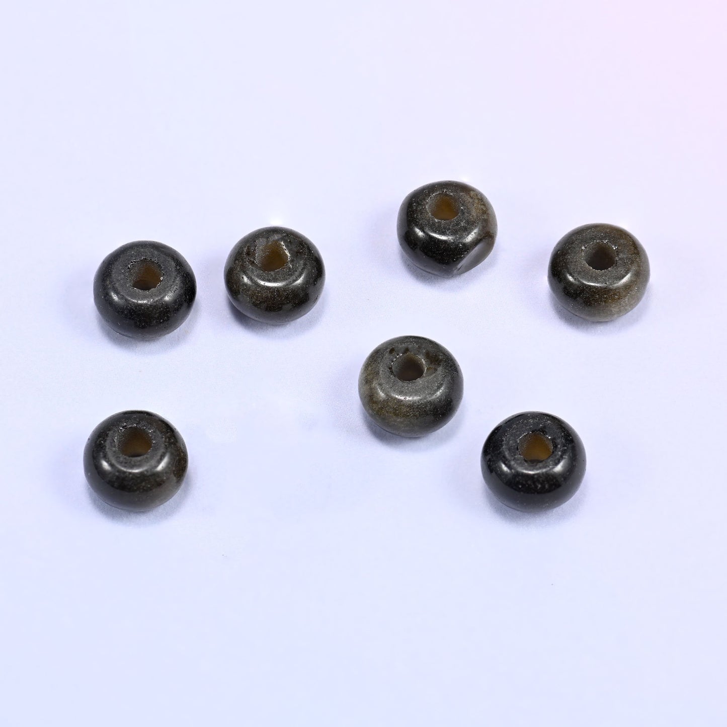 Close-up of individual loose Gold Obsidian beads laid out on a white surface, showing the large center-drilled hole and golden shimmer of each bead.