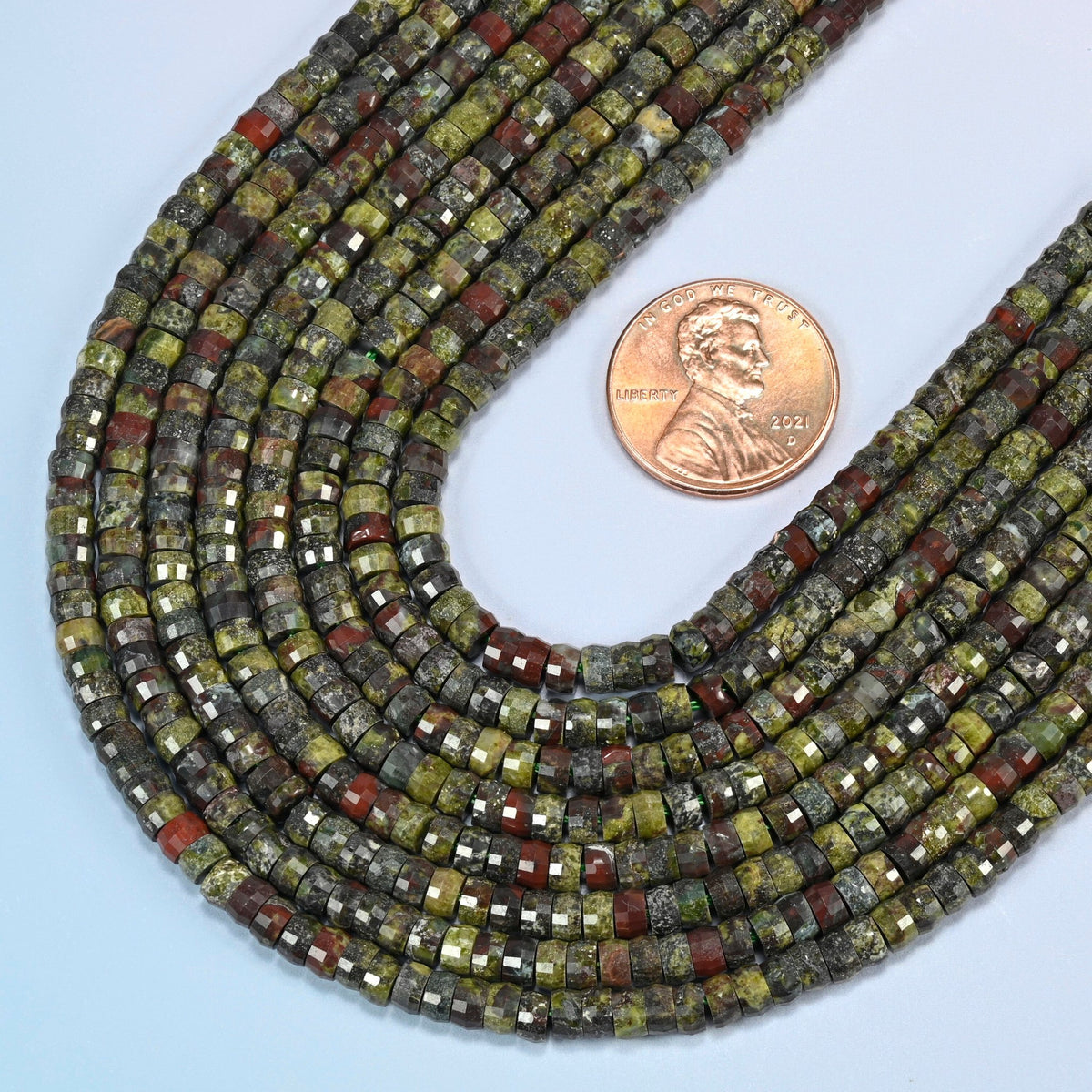 Close view of dragon blood stone faceted heishi disc strands curving beside a US penny for scale