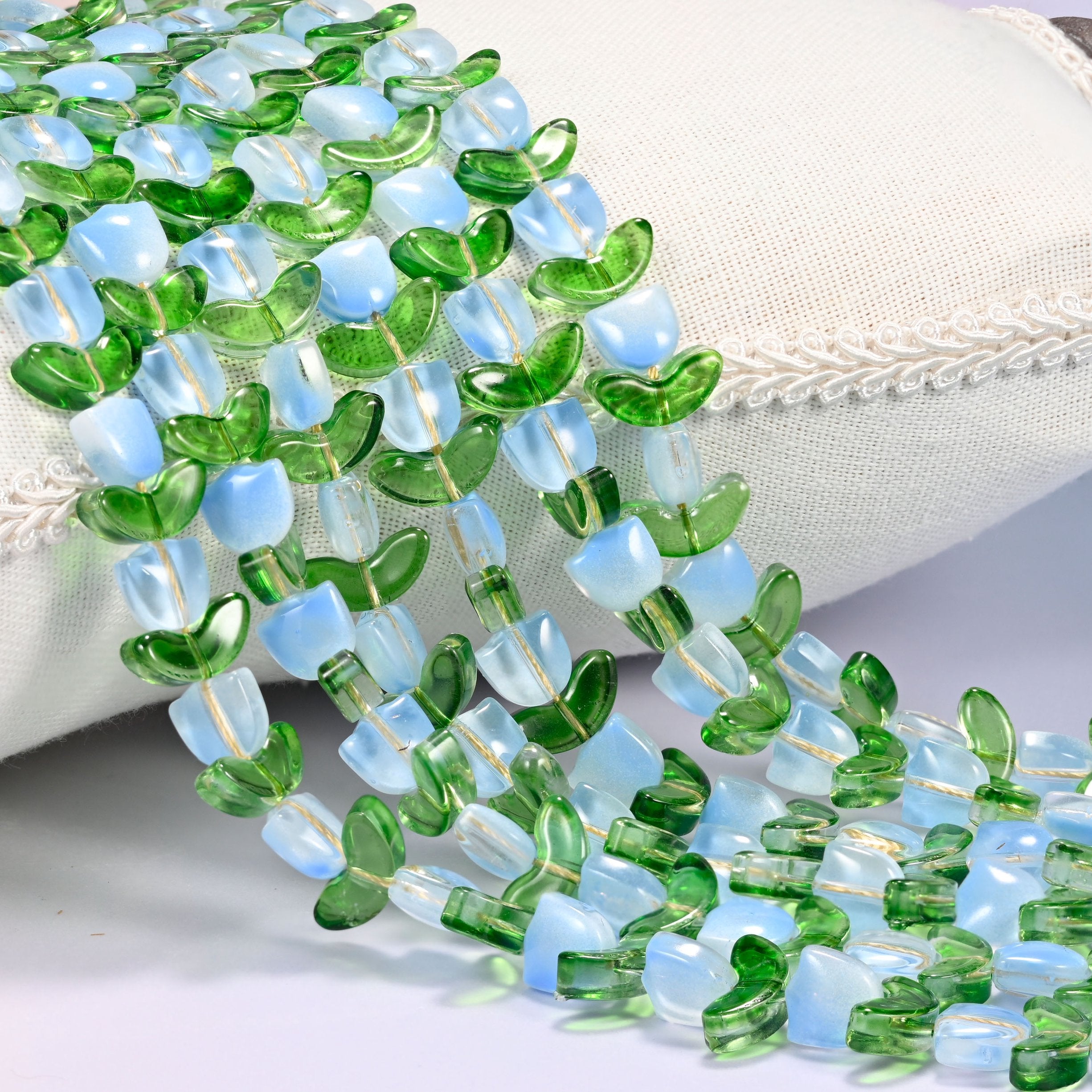 Close-up of light blue opal-toned tulip flower and green leaf glass beads cascading diagonally over a cream woven pillow with decorative trim.