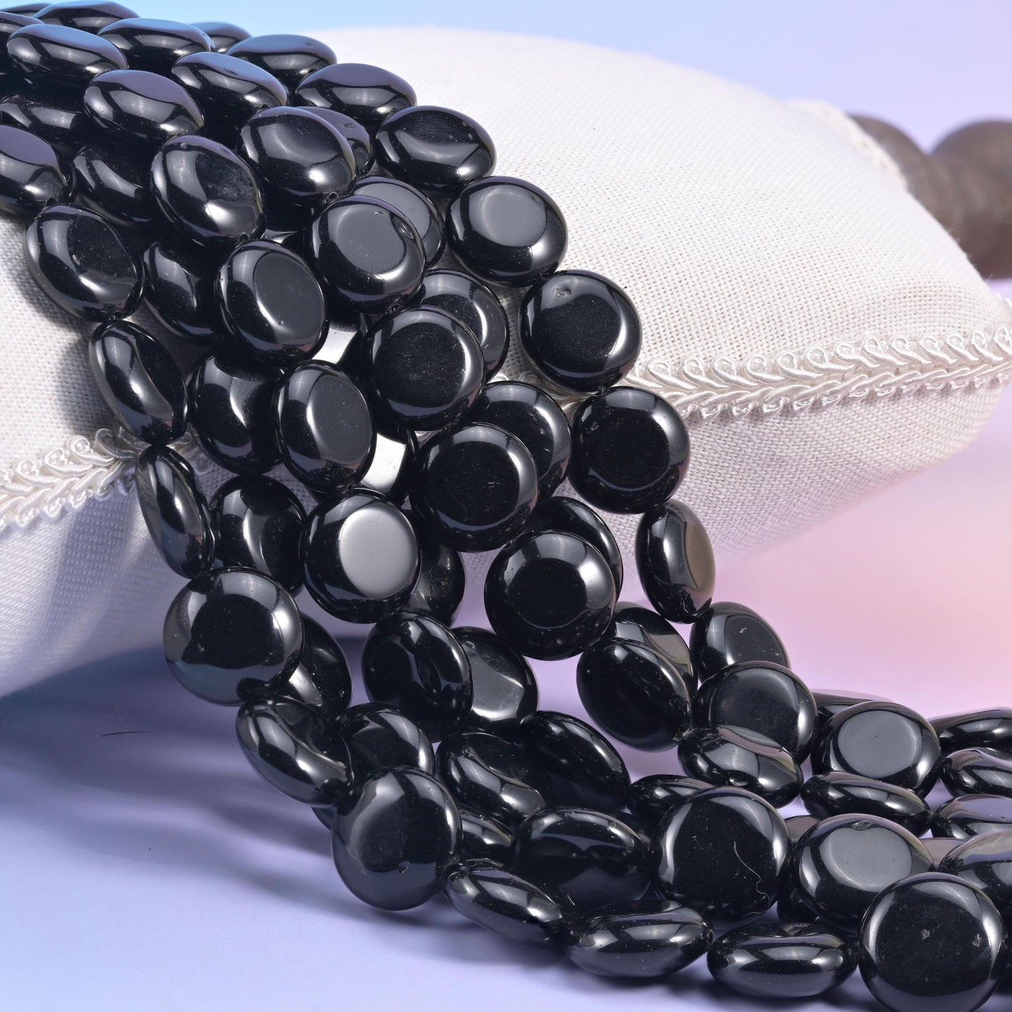Glossy black obsidian coin bead strands spilling over a white linen cushion, mirror-polished discs catching bright highlights