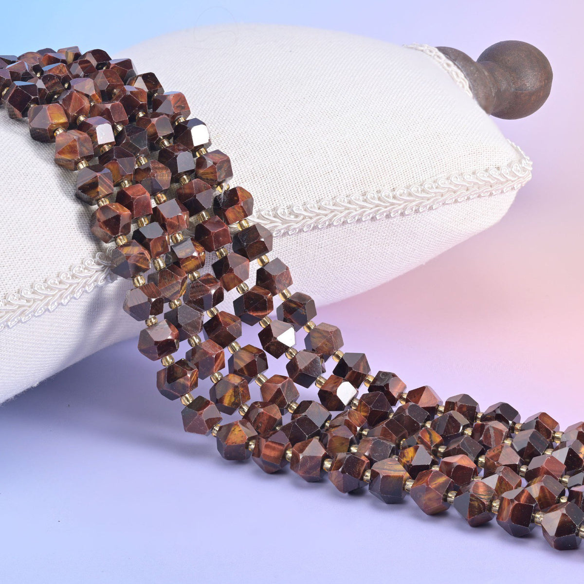 Faceted 8 sides diamond rhombus beads in dyed red tiger eye draped over a white cushion, banded red brown sheen.