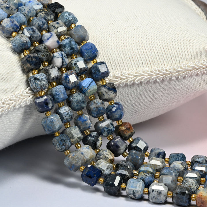 Sunset dumortierite faceted cube beads draped over a white linen cushion with braided trim, speckled blue and gray cubes with gold glass spacers