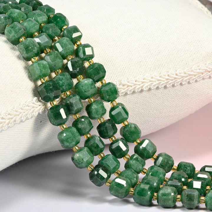 Natural green jade faceted cube beads in mottled deep green, strands draped over a white linen cushion with gold glass seed bead spacers