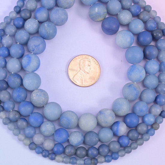 Denim blue rounds finished matte all over, so the surface reads soft and chalky rather than shiny. Pale grey-blue clouding drifts through the darker stone and gives the whole strand a faded