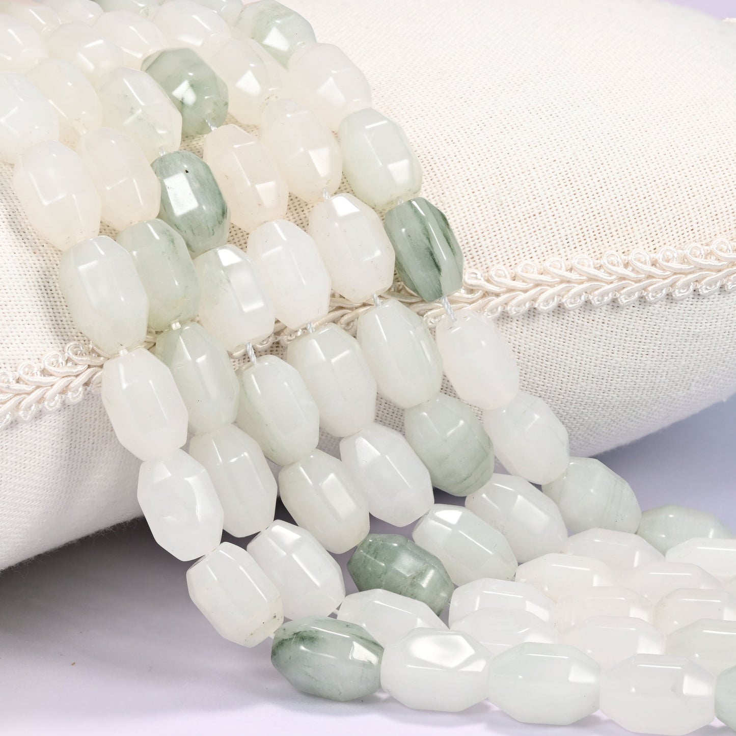 Close-up of multiple strands of white and sage green faceted Tianshan Jade barrel beads cascading diagonally over a cream woven pillow with decorative trim.