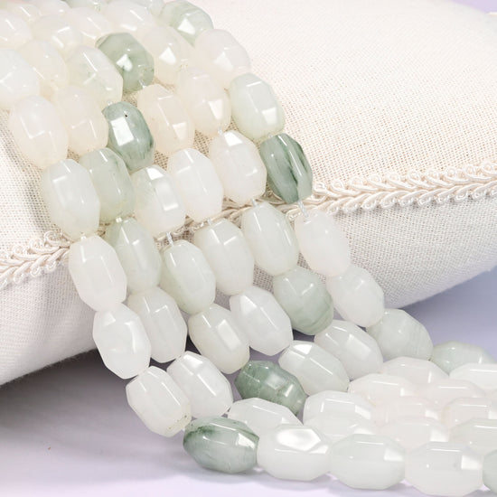 Close-up of multiple strands of white and sage green faceted Tianshan Jade barrel beads cascading diagonally over a cream woven pillow with decorative trim.