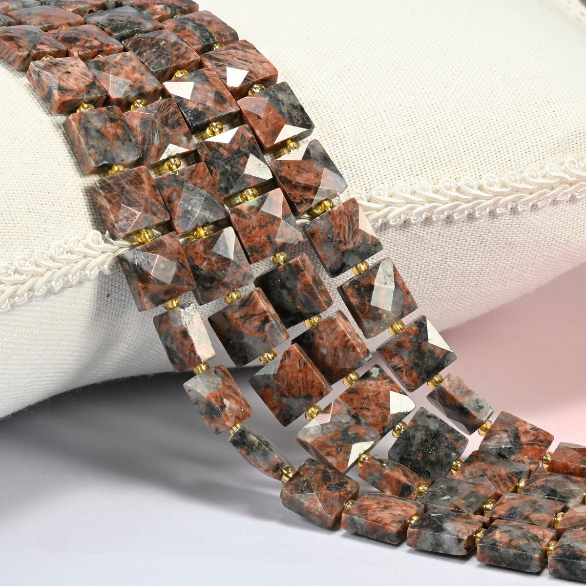 Natural red sesame jasper faceted flat square beads 10x10mm with small gold seed bead spacers, several strands draped over a white cushion