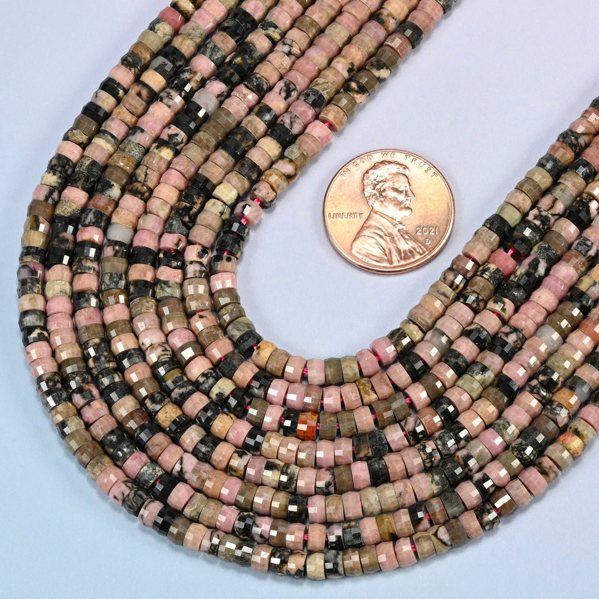 Close view of rhodonite faceted heishi disc strands curving beside a US penny for scale