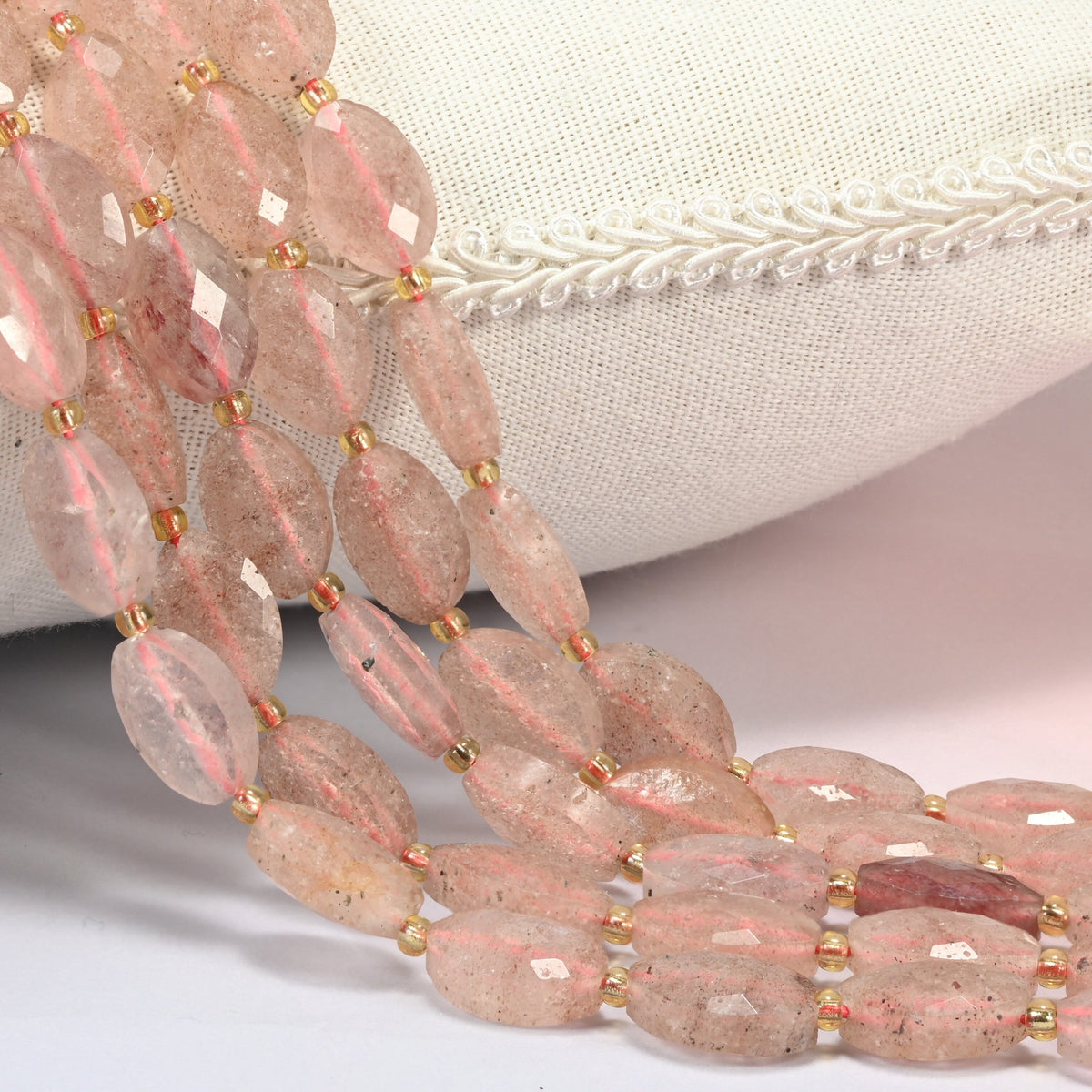 Strawberry quartz faceted flat oval beads 9x14mm with small gold seed bead spacers, several strands draped over a white cushion