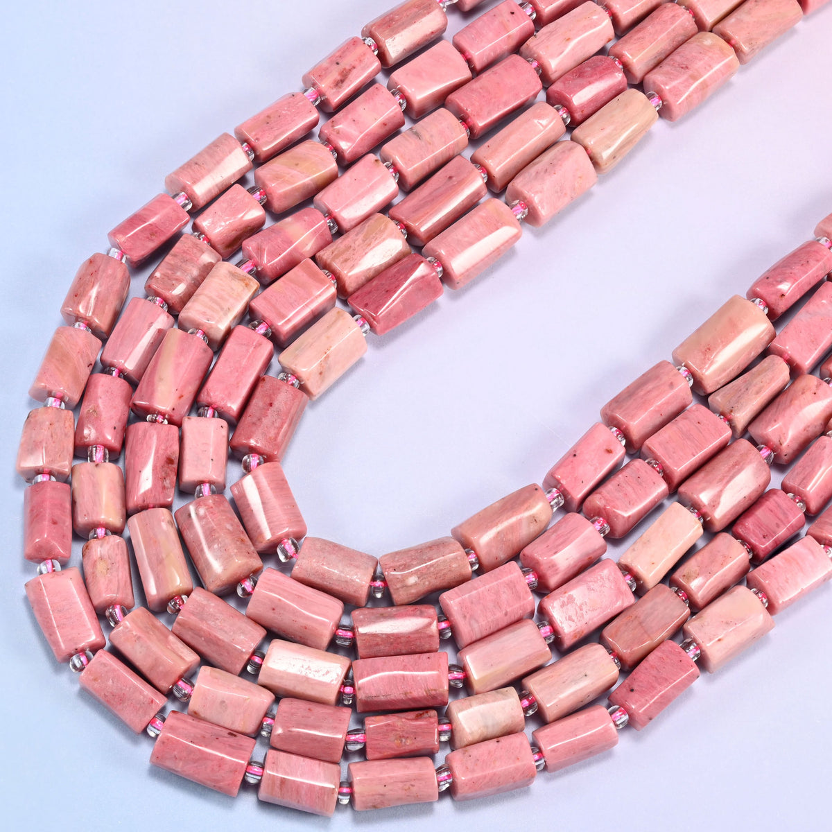 Pink Rhodonite faceted tube beads curving in a wide U across the frame, dusty rose shading into warm salmon and cream