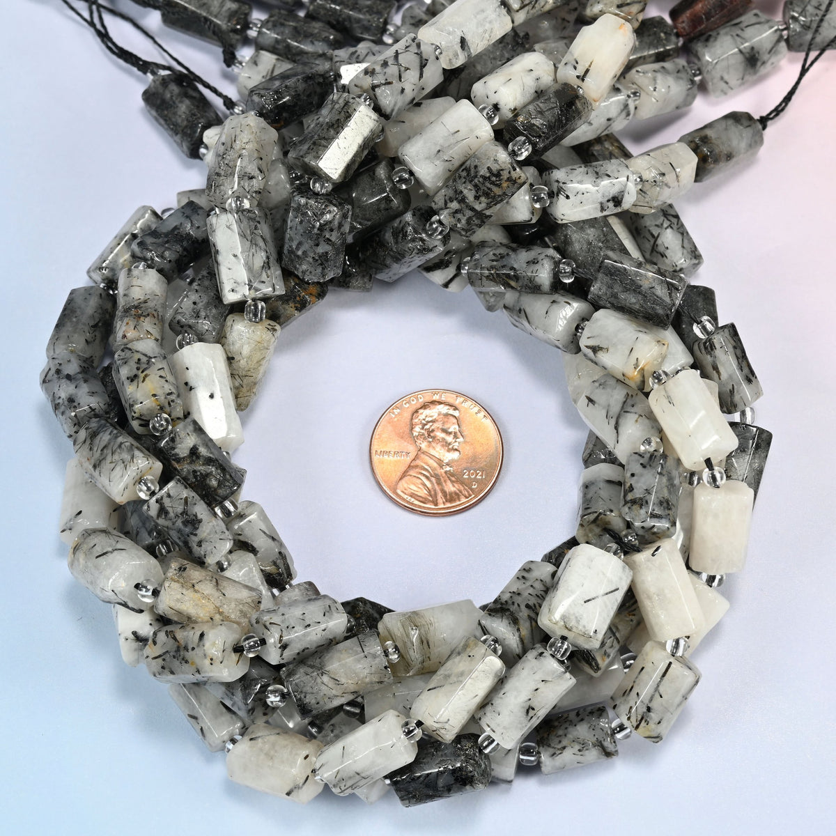 Black rutilated quartz faceted tube beads 7x11-8x12mm coiled in a loop around a US penny for scale, close-up of the faceted tube shape
