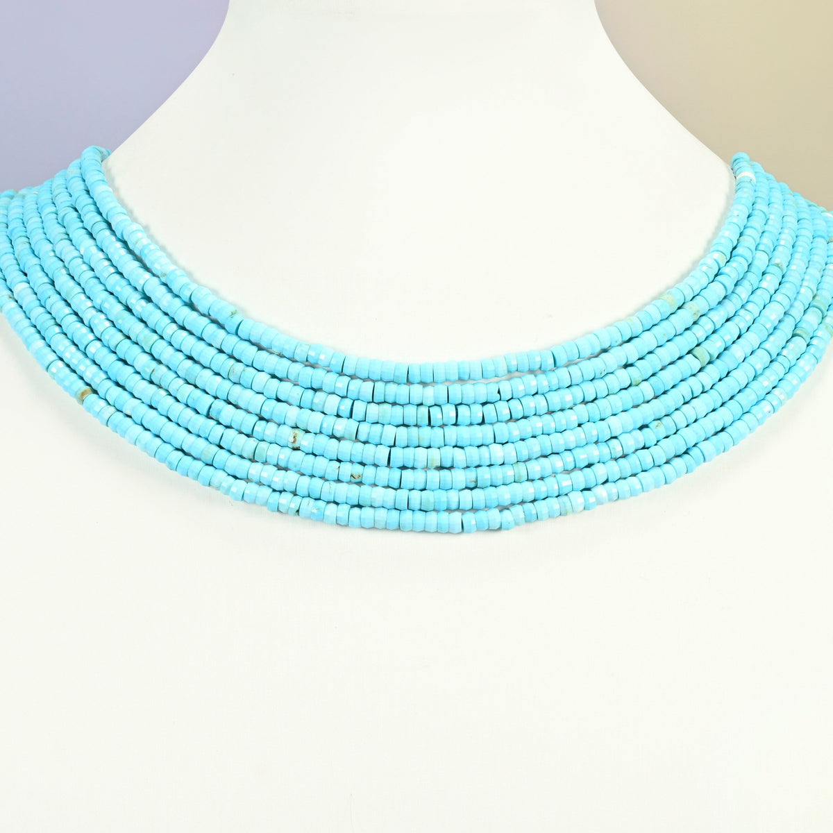 Dyed blue howlite faceted heishi disc strands arranged as a layered collar on a white display bust, turquoise blue beads across the neck