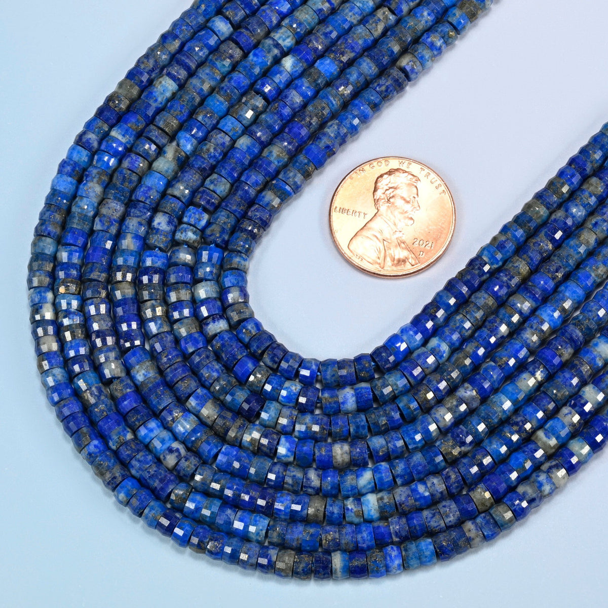 Close view of lapis faceted heishi disc strands curving beside a US penny for scale