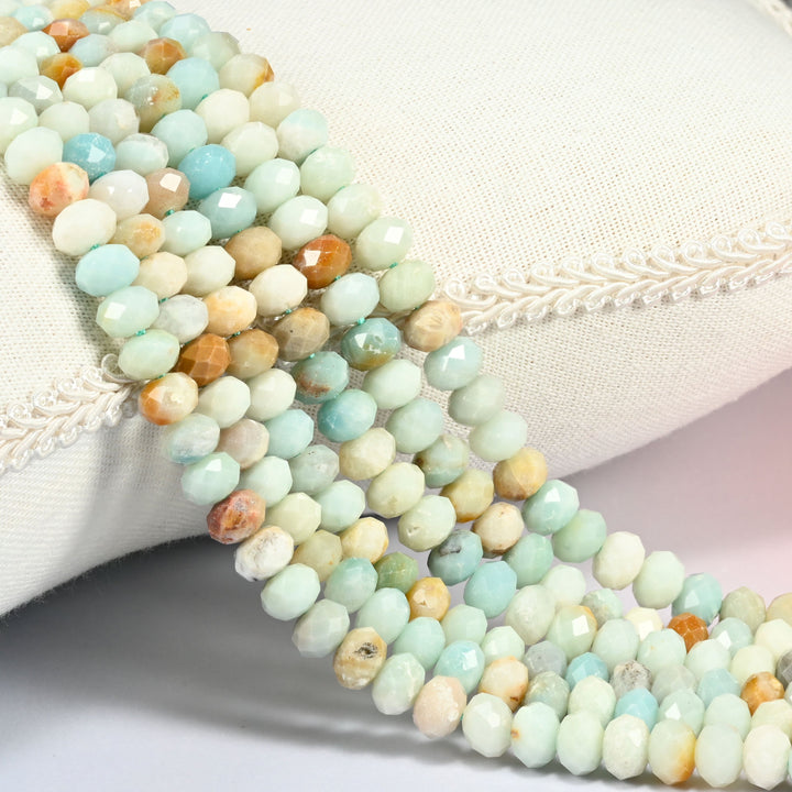 Multi-color amazonite faceted rondelle beads in mint green, cream and light brown mixed, three strands draped over a cream linen cushion with braided trim
