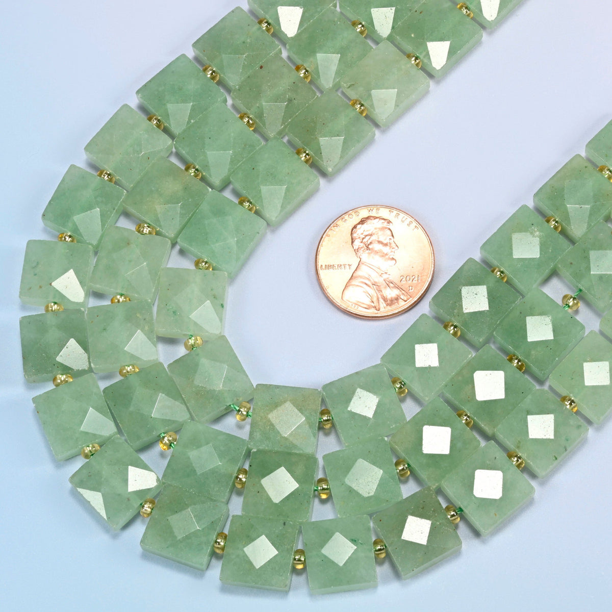 Close view of natural green aventurine faceted flat square strands curving beside a US penny for scale