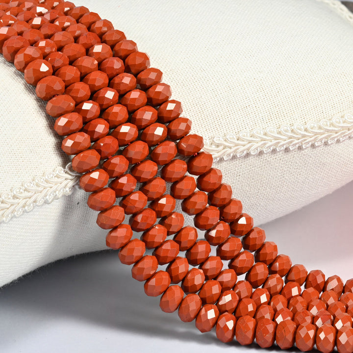 Red jasper faceted rondelle beads in deep opaque brick red, three strands draped over a cream linen cushion with braided trim