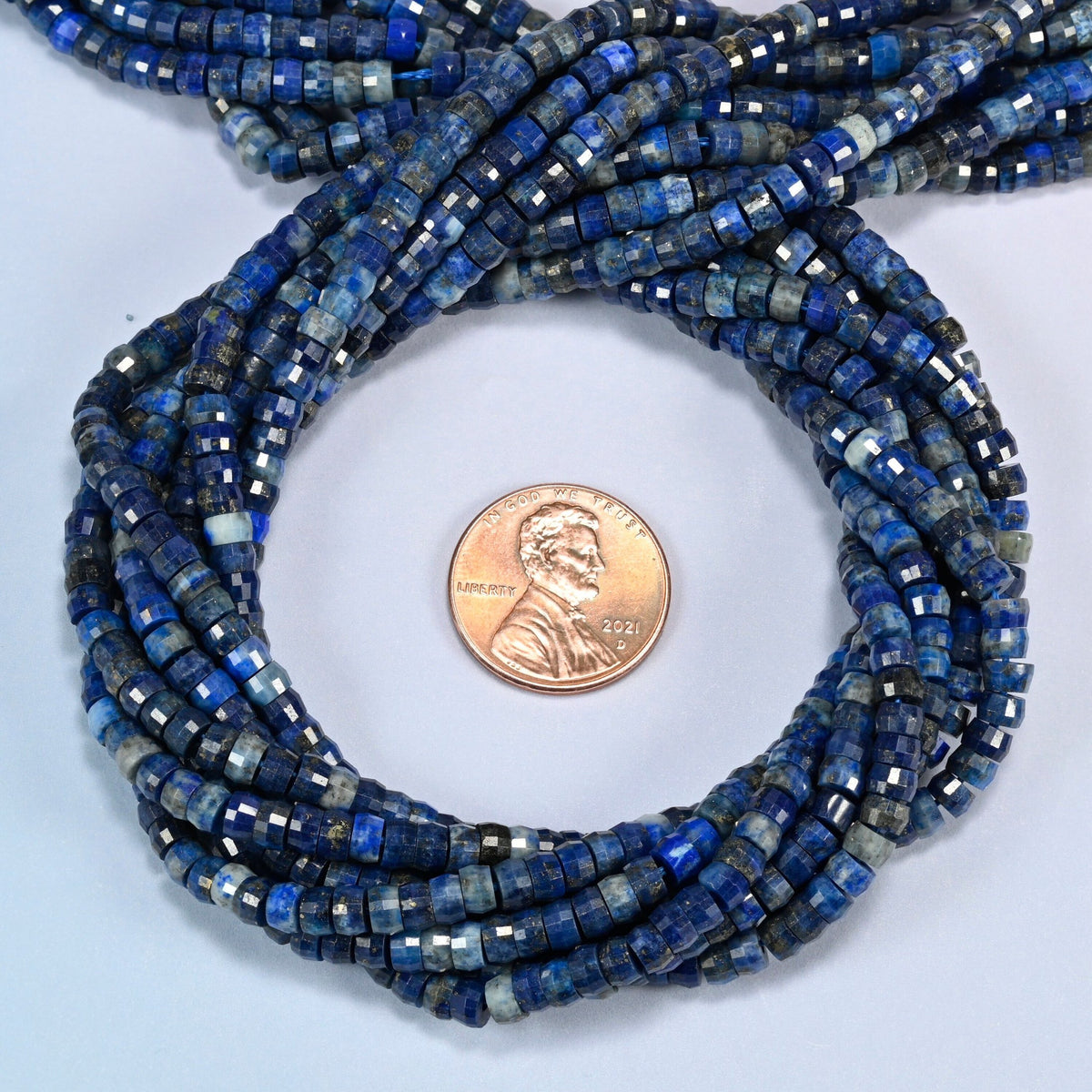 Dark blue lapis faceted heishi disc beads 2x4mm coiled in a loop around a US penny for scale, close-up of the faceted disc shape
