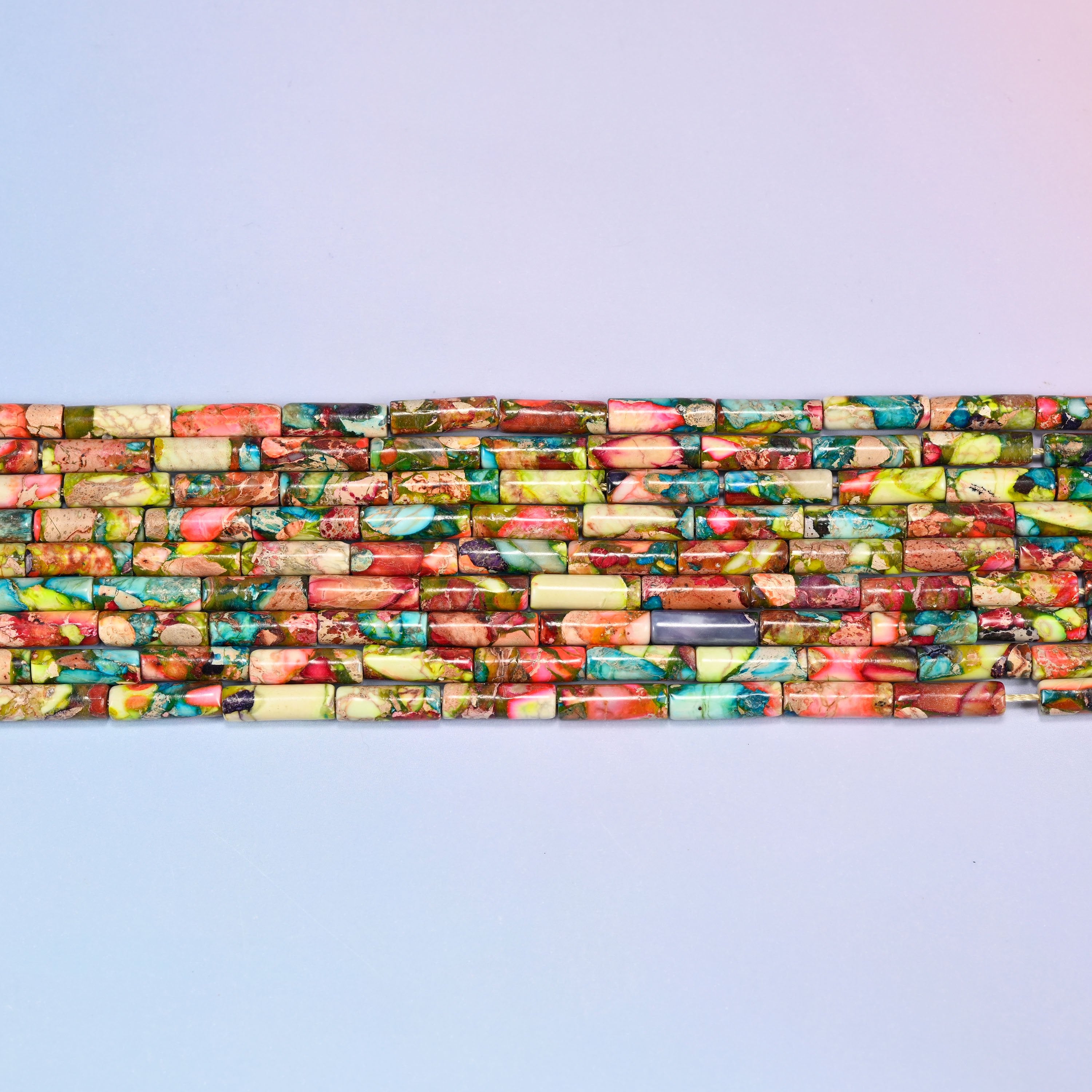 Overhead view of several rows of multicolor jasper tube beads on a light lavender surface, showing the varied stone patterns along the strands.