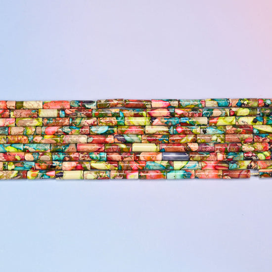 Overhead view of several rows of multicolor jasper tube beads on a light lavender surface, showing the varied stone patterns along the strands.