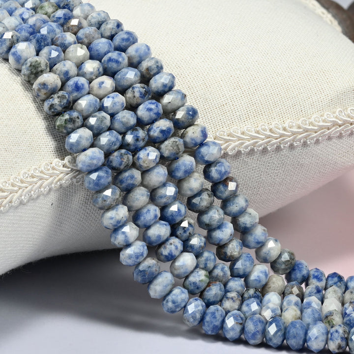 Blue spot jasper faceted rondelle beads in denim blue speckled with white and cream, three strands draped over a cream linen cushion with braided trim