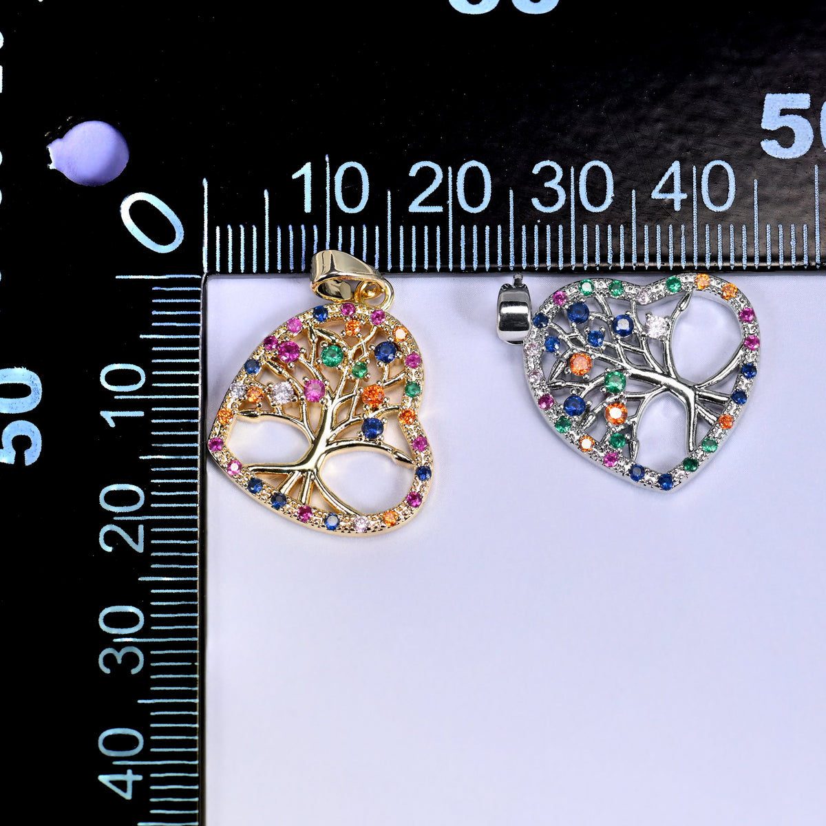 Gold and silver heart tree of life zirconia charms on a black millimetre ruler, shown together for size reference.