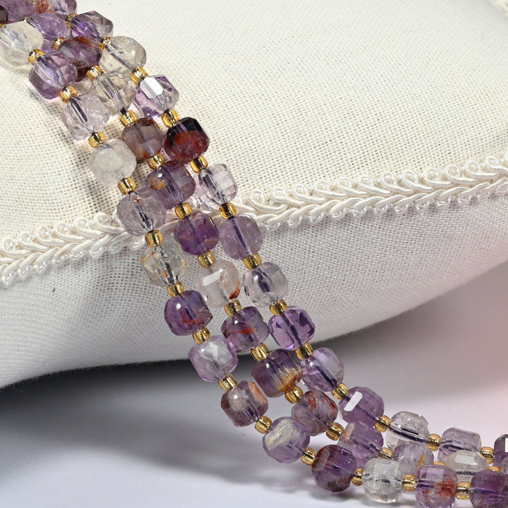 Natural Super 7 faceted cube beads with gold glass seed bead spacers, strands draped over a white linen cushion with braided trim