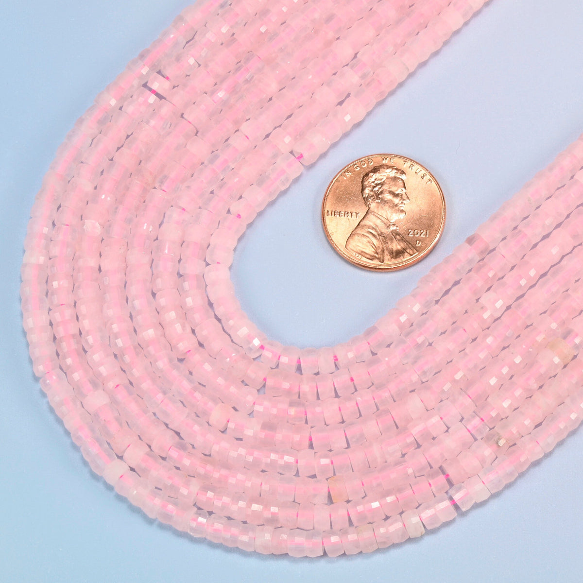 Close view of rose quartz faceted heishi disc strands curving beside a US penny for scale