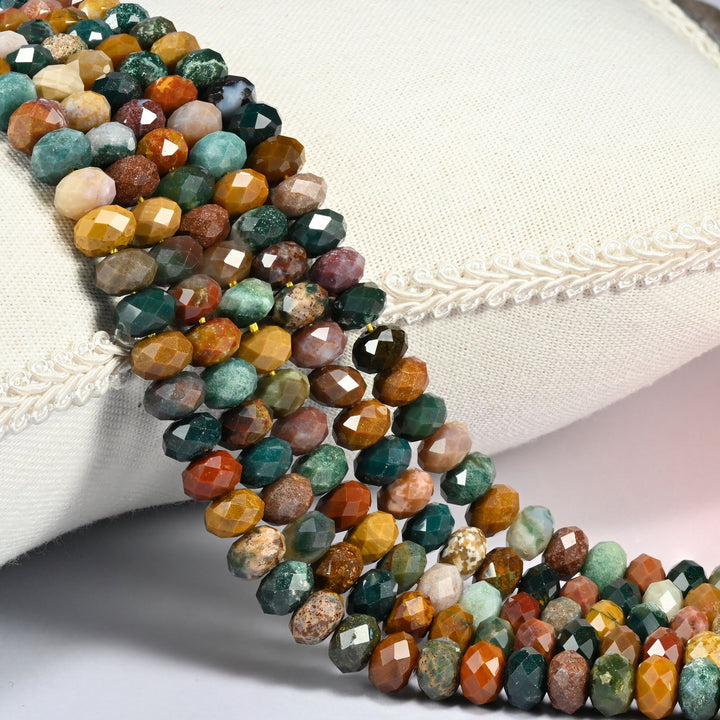 Ocean Agate faceted rondelle beads draped over a cream fabric cushion, teal and deep green against orange-brown, golden brown and light tan across the 4x6mm and 5x8mm strands.