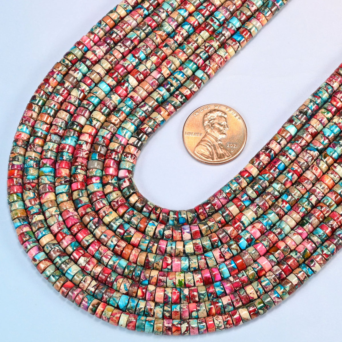 Close view of dyed pink blue sea sediment jasper heishi disc strands curving beside a US penny for scale