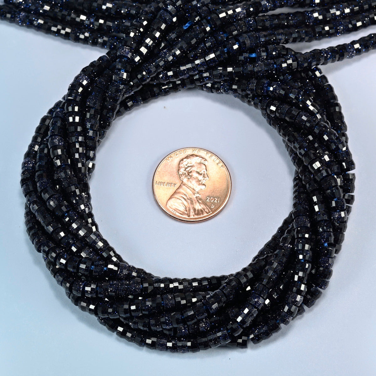 Blue sandstone faceted heishi disc beads 2x4mm coiled in a loop around a US penny for scale, close-up of the faceted disc shape