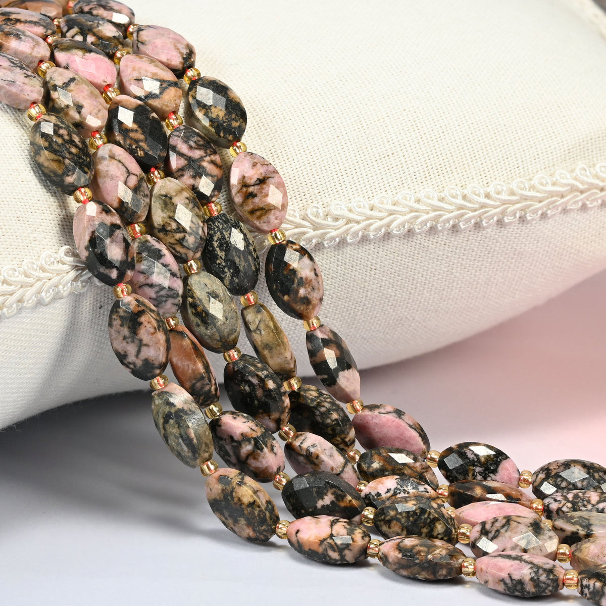 Rhodonite faceted flat oval beads 9x14mm with small gold seed bead spacers, several strands draped over a white cushion