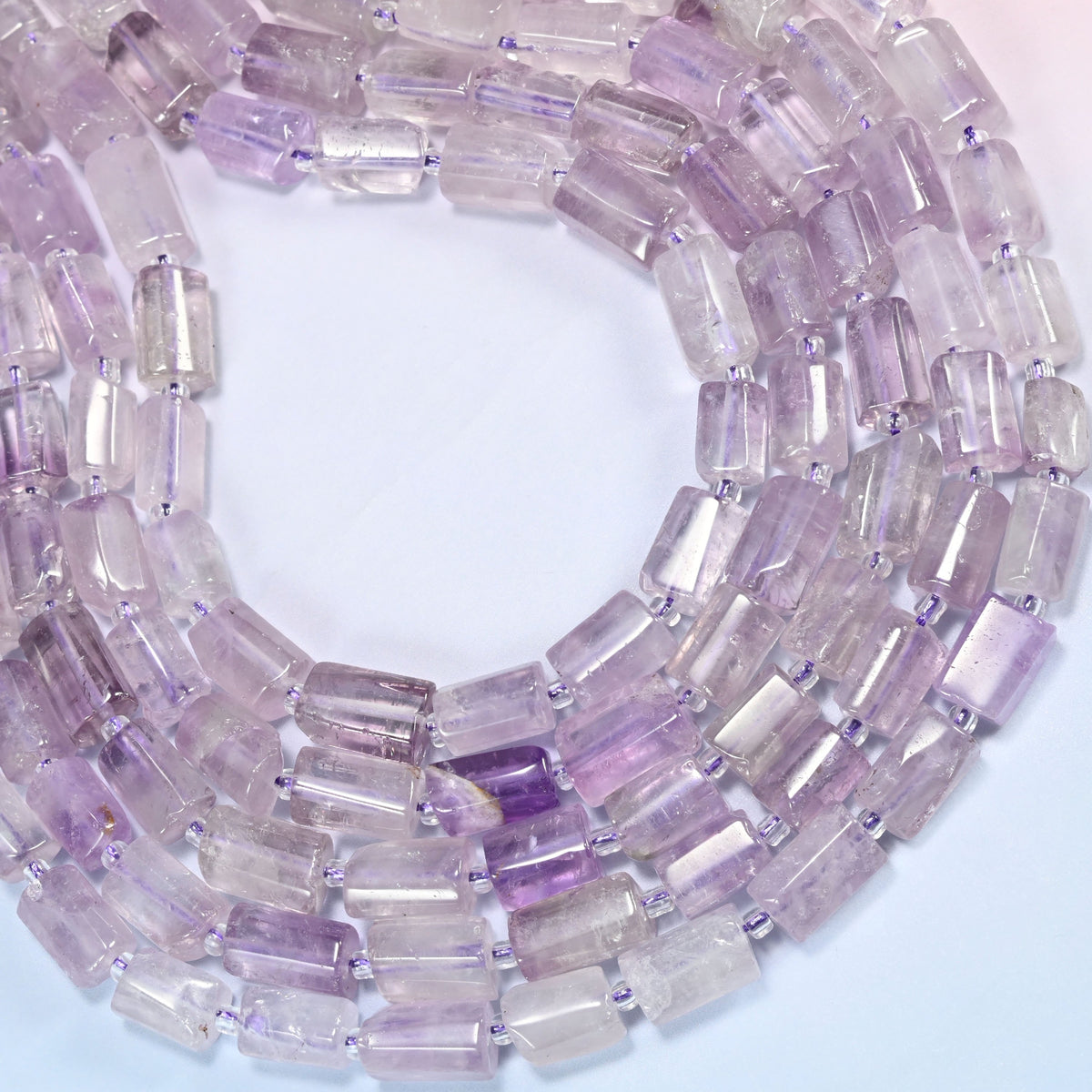 Lavender amethyst faceted tube beads wound into an open coil on a soft blue backdrop, light violet and clear tubes side by side.