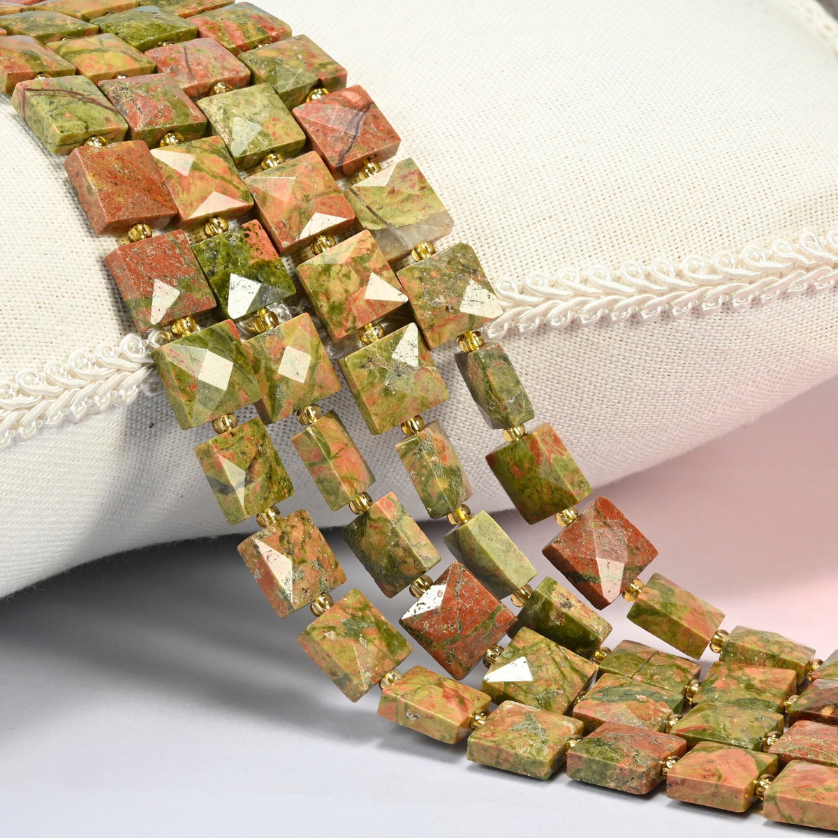 Natural unakite faceted flat square beads 10x10mm with small gold seed bead spacers, several strands draped over a white cushion