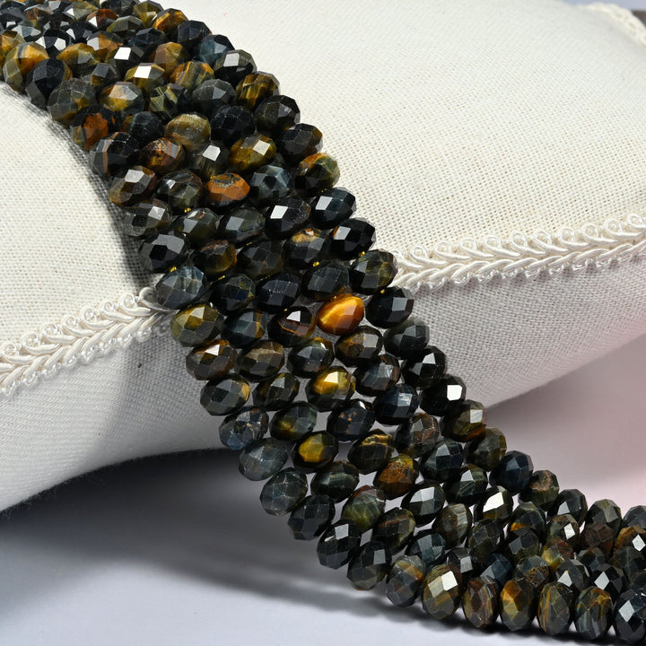 Iron tiger eye faceted rondelle beads in dark blue gray and golden brown, several strands draped over a cream cushion with braided trim