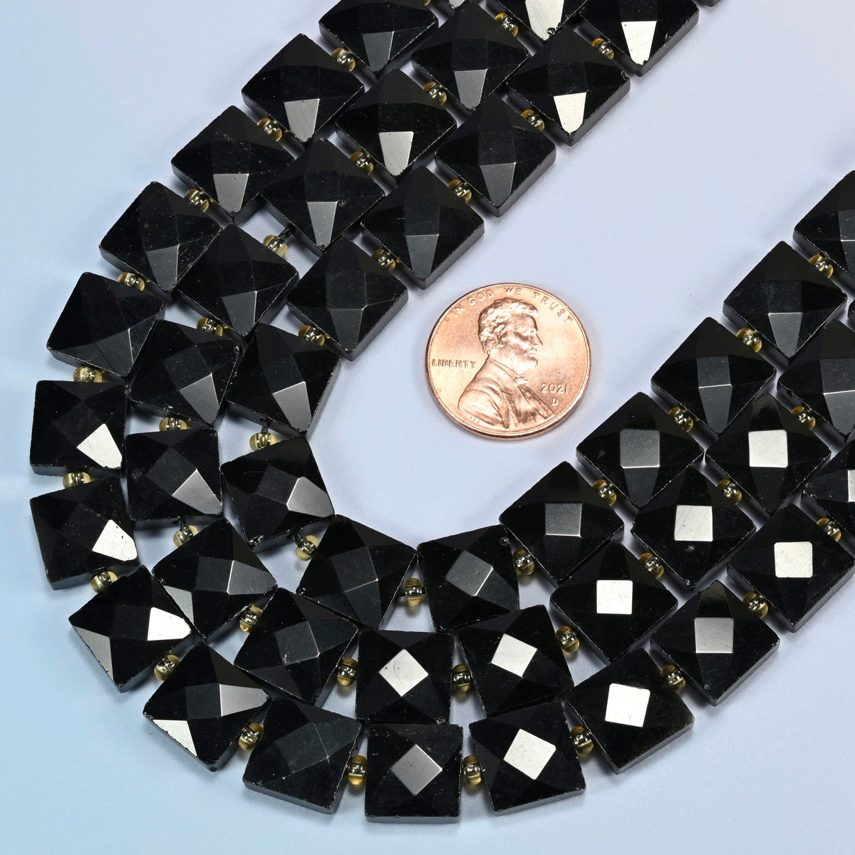 Close view of natural black onyx faceted flat square strands curving beside a US penny for scale