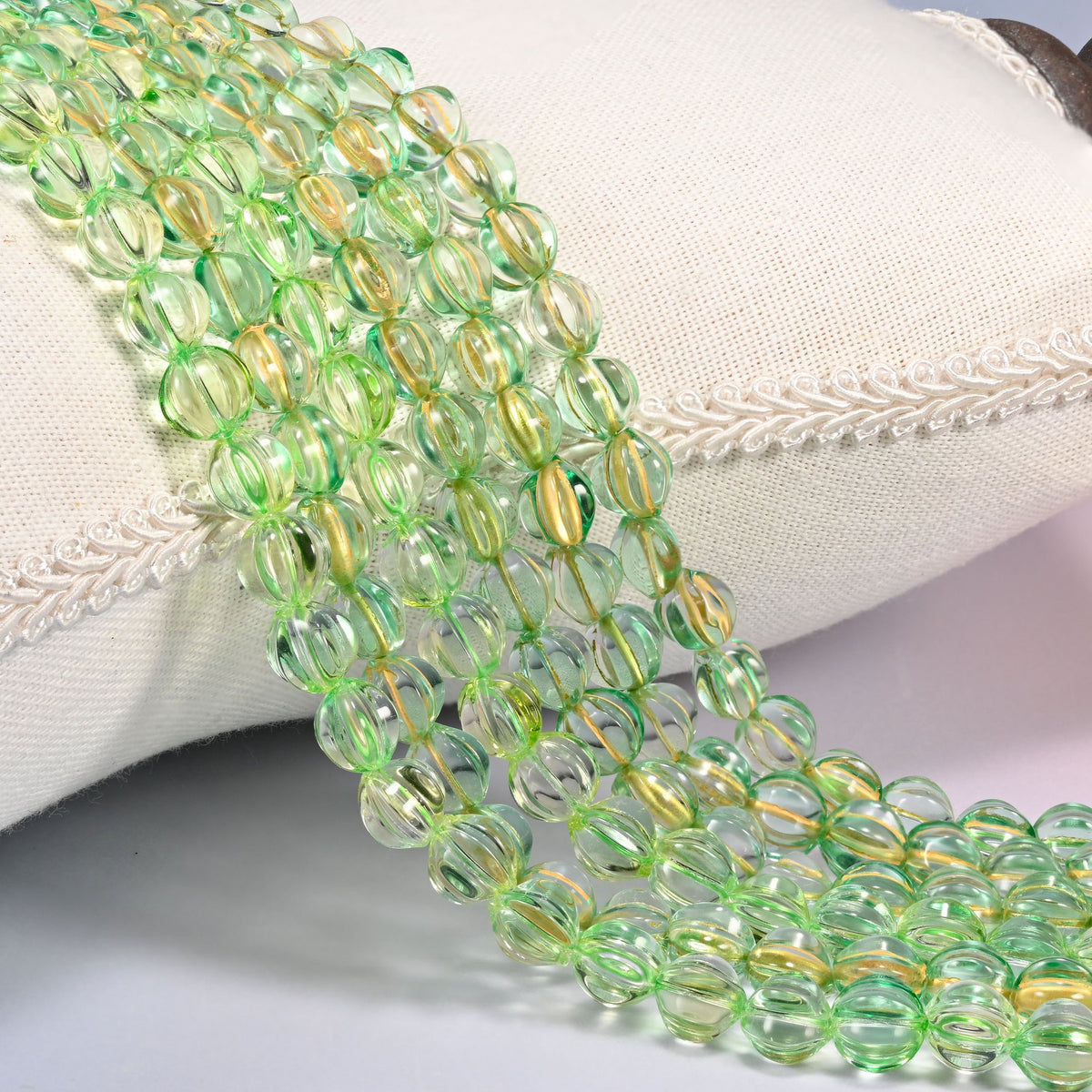 Green gold pumpkin glass beads, 8mm fluted melon rounds, several strands draped over a white cushion.