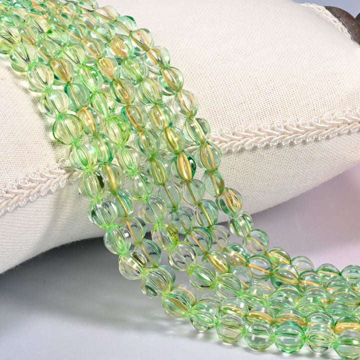 Green gold pumpkin glass beads, 8mm fluted melon rounds, several strands draped over a white cushion.