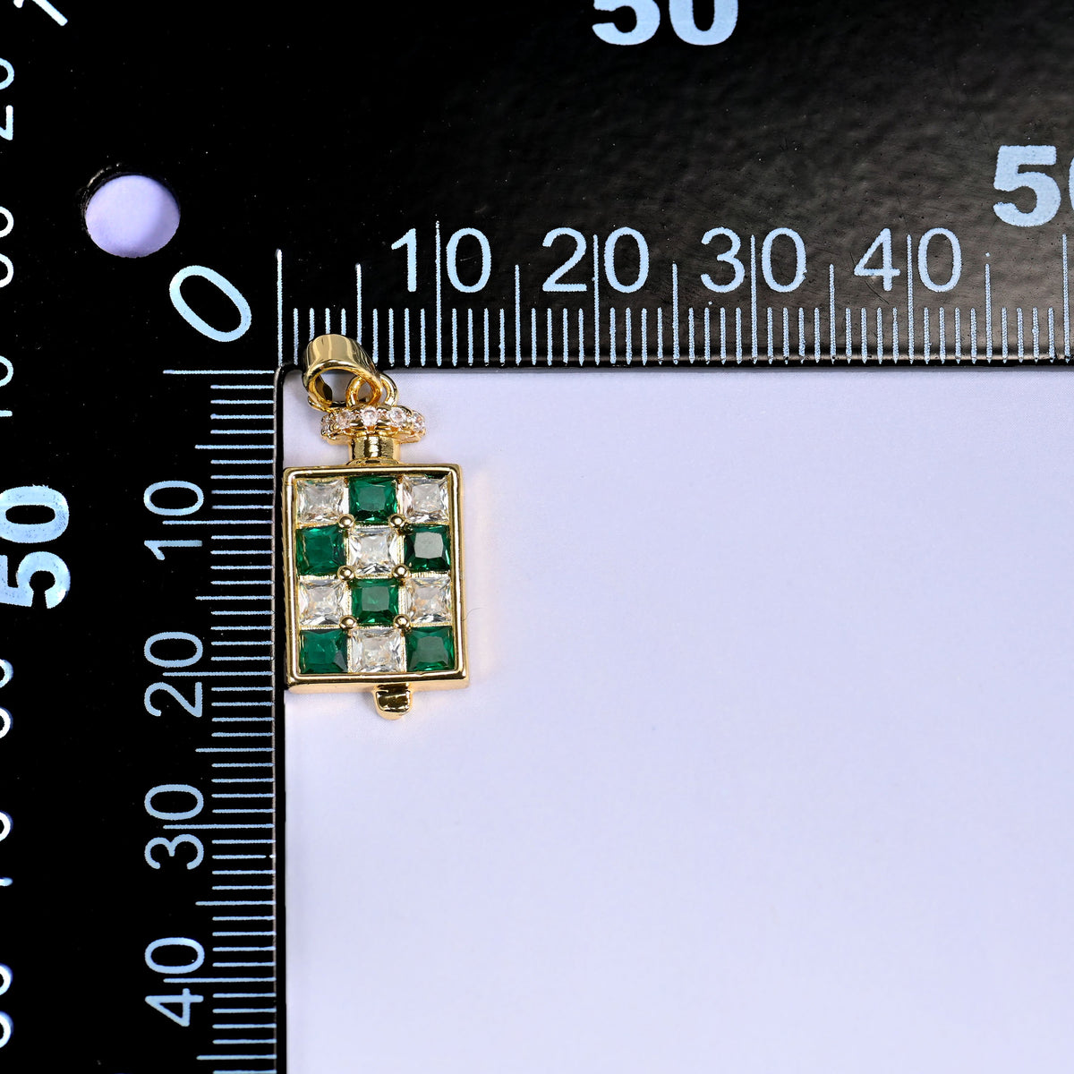 Perfume bottle zirconia charm against a black millimetre ruler, the green pendant lined up at the zero mark for size reference.