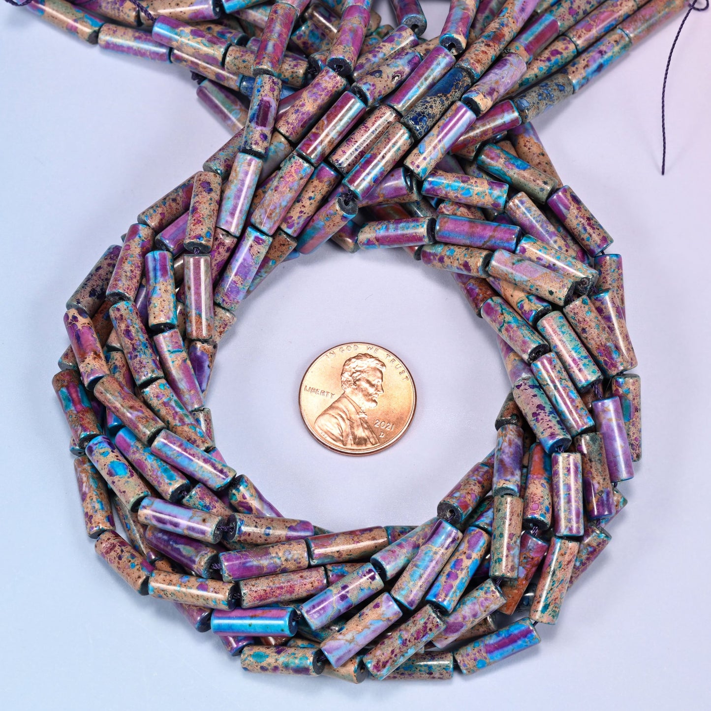 Bundled coil of purple, magenta, and turquoise jasper tube beads on a lavender background with a penny placed at the center for scale.