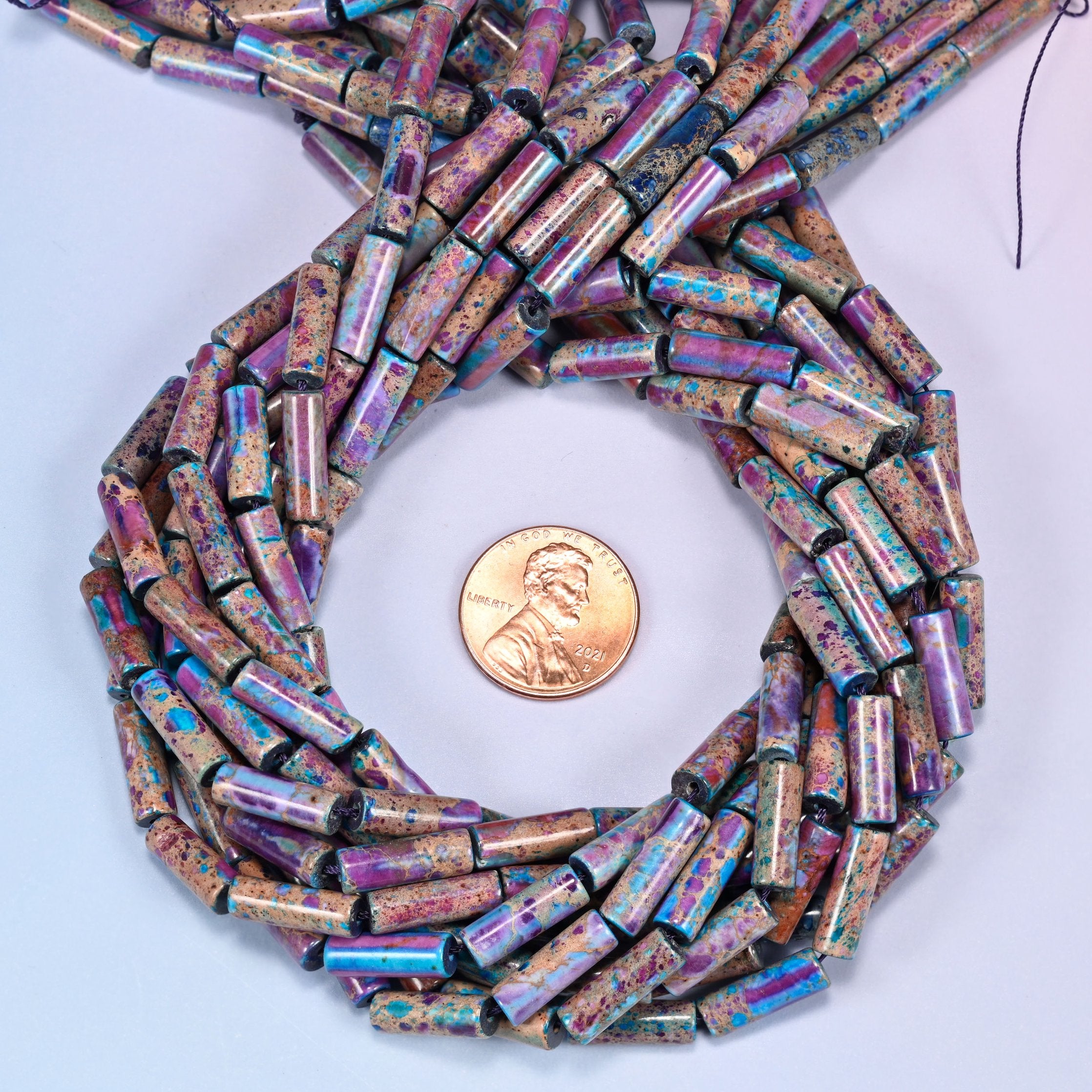 Bundled coil of purple, magenta, and turquoise jasper tube beads on a lavender background with a penny placed at the center for scale.
