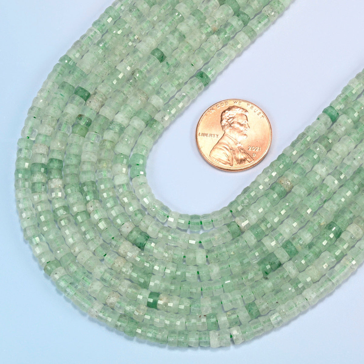Close view of green strawberry quartz faceted heishi disc strands curving beside a US penny for scale