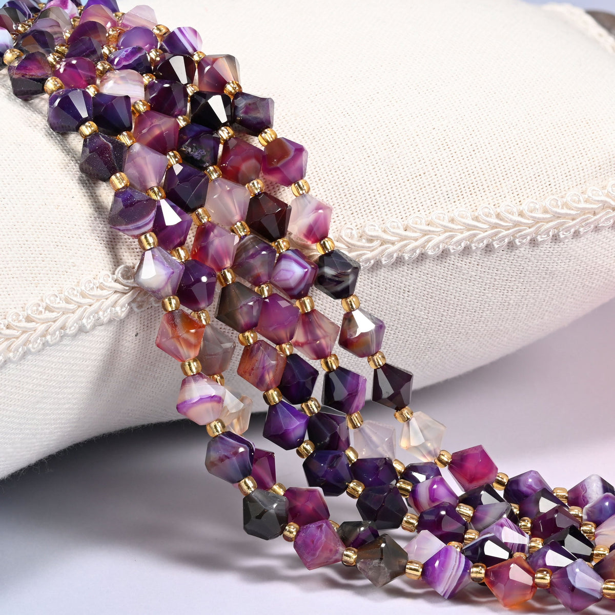 Dyed Purple Strip Agate faceted bicone beads, 8mm, draped diagonally over a white fabric cushion showing the purple color with natural banded striping.