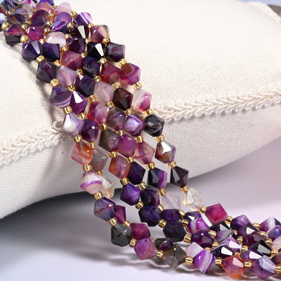 Dyed Purple Strip Agate faceted bicone beads, 8mm, draped diagonally over a white fabric cushion showing the purple color with natural banded striping.