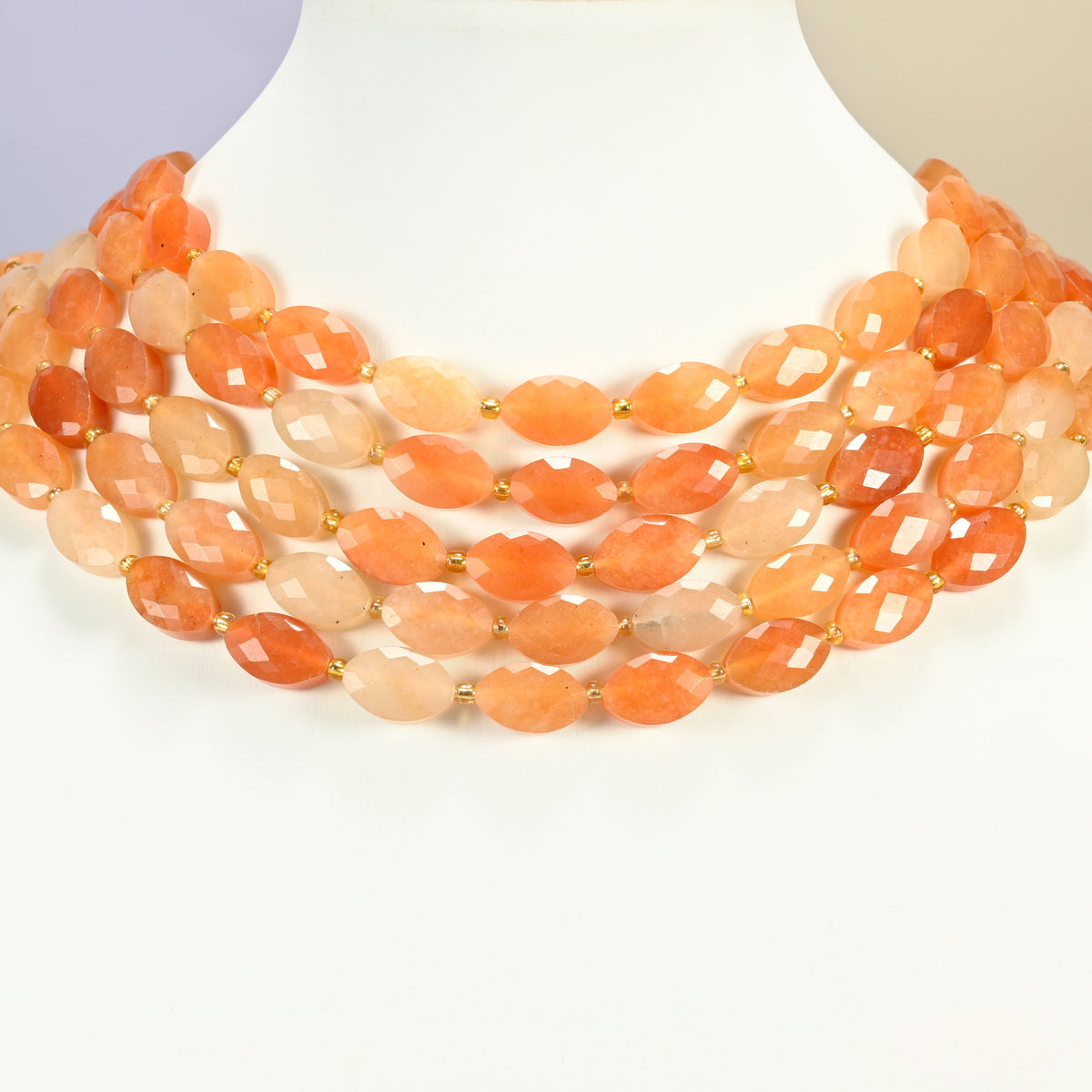 Red aventurine faceted flat oval strands arranged as a layered collar on a white display bust, peach and orange beads across the neck